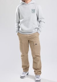 Grå hoodie med framficka och tryckt logotyp; kombinerad med beige cargo-byxor med sidofickor. Vita sneakers med grå detaljer.