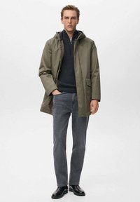 Chaqueta impermeable de color verde oliva con capucha, que presenta un cierre de cremallera y dos bolsillos frontales, llevada sobre un suéter de punto azul marino y unos jeans grises.