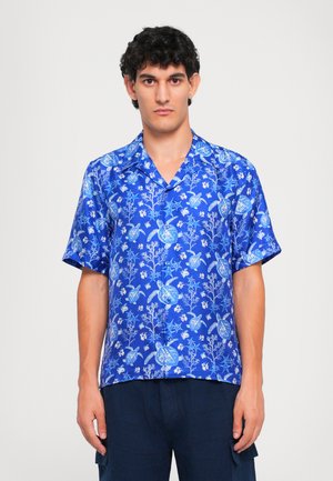 Young man wearing a bright blue short-sleeve shirt with a sea turtle and coral pattern, paired with dark blue pants.