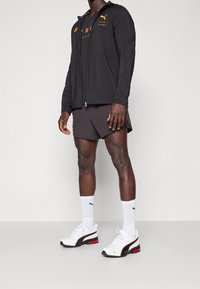 Chaqueta negra con cremallera y acentos en amarillo y rojo, combinada con una camiseta negra, pantalones cortos negros y zapatillas blancas con detalles en rojo y negro.