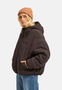 Femme portant un bonnet marron et une veste matelassée marron foncé oversize à capuche, les mains dans les poches, associée à un jean bleu.
