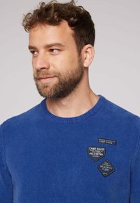 Maglione blu reale con scollo rotondo a costine, tessuto strutturato e tre toppe nere in tessuto con testo e loghi.