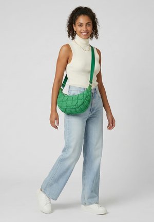 Frau mit lockigem Haar, die einen weißen ärmellosen Rollkragenpullover und hellblaue Jeans trägt, eine grüne gesteppte Umhängetasche mit sich führt und lächelt.