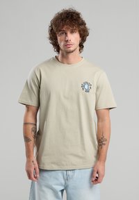 Camiseta de algodón beige claro con un diseño gráfico azul en el lado izquierdo del pecho, escote redondo, mangas cortas y un ajuste holgado.