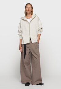 Sudadera gris claro con cremallera y capucha, de corte holgado y con puños acanalados; combinada con pantalones taupe de talle alto y pierna ancha, y zapatos planos negros.