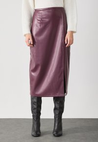 Burgunderfarget faux skinn midi skjørt med sideslitt og søm detaljer, kombinert med svarte knelengde støvler på en grå overflate.
