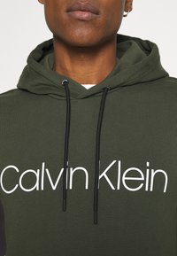 Bluza z kapturem w oliwkowej zieleni z białym napisem "Calvin Klein", z kapturem na sznurek i teksturowaną tkaniną. Zbliżenie na szczegóły przy szyi i logo.