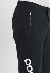 Black outdoor pants with a smooth texture, featuring a zippered pocket and white logo detail on the side.