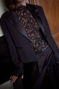 Schwarzer Blazer mit Satinrevers über einem schwarzen Spitzentop, das mit Pailletten verziert ist. Flared schwarze Hosen und ein Ledergürtel vervollständigen das Outfit.