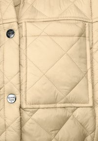 Prošívaná béžová bunda z látky s velkou přední kapsou a logovací knoflíkem na boku, která zdůrazňuje měkkou texturu a prošívaný vzor.