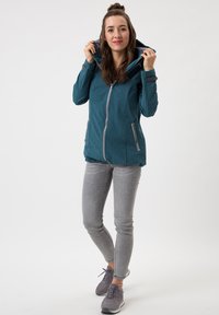 Türkise Zip-Jacke mit Kapuze und Seitentaschen, getragen über grauen Slim-Fit-Jeans. Der Stoff hat eine strukturierte Oberfläche und einen leichten Glanz.