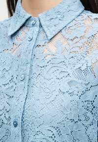 Ανοιχτό μπλε δαντέλα πουκάμισο με λουλουδένια σχέδια, κουμπωμένο μέχρι το γιακά, που δείχνει την περίπλοκη υφή και το σχέδιο του υφάσματος.