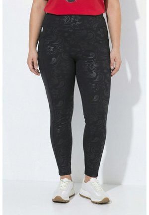 Person trägt schwarze Leggings mit dezentem Blumenmuster, rotes Oberteil und weiße Turnschuhe und steht vor einem weißen Hintergrund.