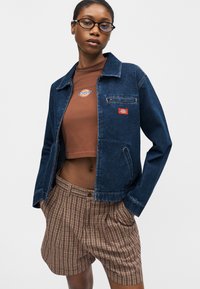 Γυναίκα με γυαλιά, τζιν μπουφάν πάνω από καφέ κοντό τοπ και καρό σορτς, στέκεται με τα χέρια στις τσέπες μπροστά σε λευκό φόντο.