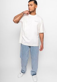 Kræmfarvet oversized t-shirt med korte ærmer og diskret "Dropsize" tekst. Sammen med lyseblå denimjeans og hvide sneakers.