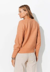 Pull à col rond, de couleur rouille orangé, en tricot doux, avec des poignets et un ourlet côtelés, et des détails tricotés décoratifs sur les épaules.