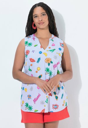 Femme aux cheveux tressés portant un chemisier sans manches coloré avec des motifs tropicaux et un short rouge, debout sur un fond blanc.