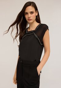 Zwarte blouse met korte mouwen, voorzien van een gestructureerde zilveren accent bij de halslijn. Gedirigeerd met hoge taille zwarte broeken.