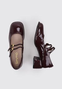 Zapatos de charol burdeos con punta cuadrada, dos correas en el tobillo y un tacón grueso. Textura suave y pequeños acentos dorados.