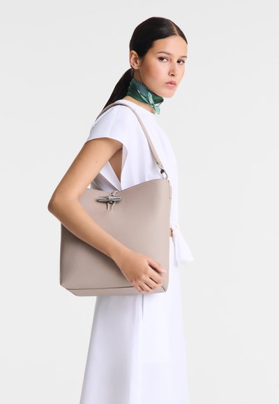 Beige strukturierte Umhängetasche mit einem silbernen Akzentbeschlag, gehalten von einer Person, die ein weißes Kleidungsstück und einen grünen Schal trägt.