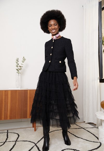 Veste noire texturée avec des boutons dorés, associée à une jupe en tulle noire à volants. Bottines noires. Le mannequin porte un foulard coloré.