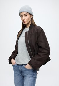 Bruine bomberjack met ritssluiting, elastische manchetten, gecombineerd met een lichtgrijze trui en lichtblauwe jeans. Lichtgrijze muts gedragen.
