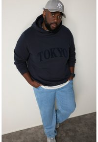Hoodie navy con la scritta "TOKYO", jeans azzurri chiari, cappellino grigio e sneakers grigie. Stile streetwear casual con una vestibilità rilassata.