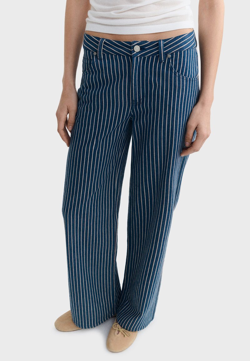 Femme portant un pantalon en denim bleu taille haute à jambes larges avec des rayures blanches verticales et des ballerines beige, vue de la taille aux pieds.