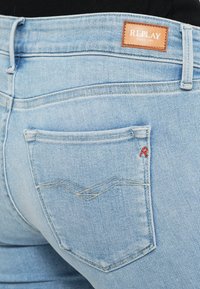 Un jean en denim bleu clair présente une poche cousue avec un motif en zigzag et un accent "R" rouge. Une étiquette de marque en cuir beige est placée à la taille.