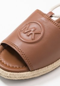 Sandale en cuir marron à enfiler avec bout ouvert, logo en relief et semelle en jute tressé. Présente des coutures visibles le long du bord supérieur.