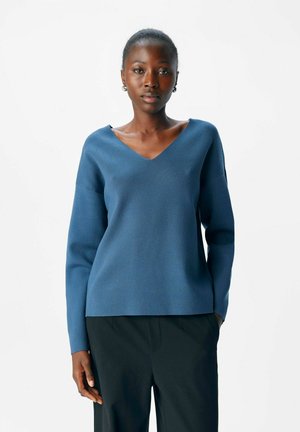 Femme à la peau foncée portant un pull bleu en V et un pantalon noir, se tenant avec une main dans sa poche contre un fond blanc uni.
