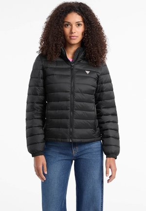 Schwarze gesteppte Jacke mit hohem Kragen, Reißverschluss und elastischen Bündchen, kombiniert mit blauen Jeans. Auffälliges Logo auf der Brust.