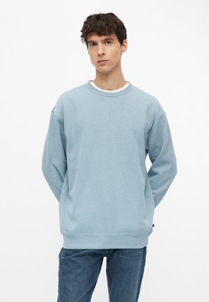 Jeune homme aux cheveux courts et bouclés porte un pull côtelé bleu clair et un jean bleu, debout, les mains derrière le dos sur fond blanc.