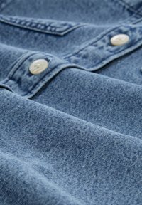 Denim stof i mellemblå med en let tekstur, der har hvide knapper og en lommeklap. Stoffet viser subtile farvevariationer.