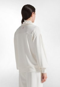 Mujer con el pelo oscuro en una coleta baja, vestida con una blusa blanca de manga larga holgada y pantalones a juego, vista desde atrás sobre un fondo blanco.