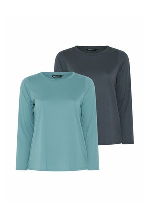 Frau mit langen dunklen Haaren, die ein dunkles Langarmshirt, schwarze Hose und weiße Turnschuhe trägt, steht vor einem schlichten hellen Hintergrund.