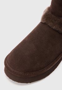 Botine din suede maro la glezna, cu un tiv moale din blană, vârf rotund și talpă din cauciuc cu detalii de cusătură vizibile pe margine.
