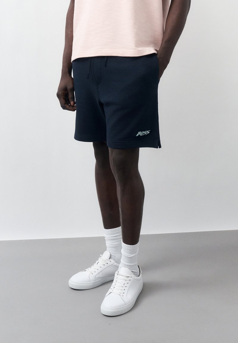 Άνδρας με μαύρα σορτς BOSS, λευκές κάλτσες και λευκά αθλητικά παπούτσια στέκεται μπροστά σε απλό γκρι και λευκό φόντο.