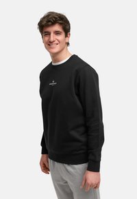 Sudadera negra con cuello redondo, puños y bajo acanalados, que presenta un logo blanco en el pecho. El modelo lleva pantalones de chándal grises.