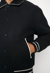 Homme portant une veste universitaire noire avec liseré blanc et poignets rayés, main partiellement dans la poche latérale, visible du cou à la taille.