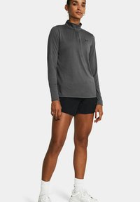 Graues Langarm-Top mit Viertelreißverschluss und glatter Textur, mit kleinem Logo; kombiniert mit schwarzen Sportshorts und weißen Sneakers.