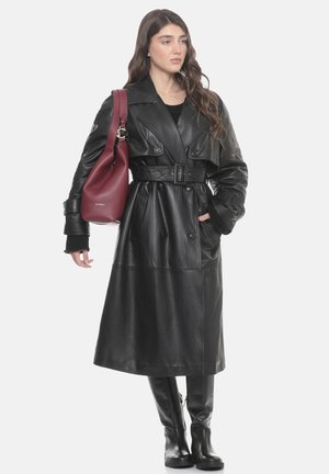 Woman with long hair wearing a belted black leather trench coat, knee-high black boots, and carrying a large red shoulder bag.