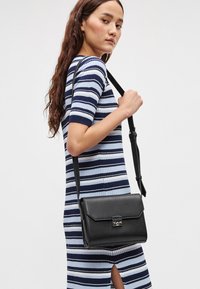 Μαύρη τσάντα ώμου με καπάκι και ασημένιο κούμπωμα, λείο υλικό, ορθογώνιο σχήμα, φορεμένη χιαστί πάνω από φόρεμα με ρίγες.