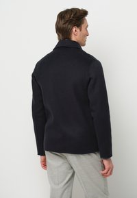 Giacca di lana blu navy con colletto a punta, spalle strutturate e una texture liscia, abbinata a pantaloni grigi chiari.