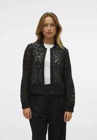 Veste en dentelle noire avec un col rond, fermeture à pression à l'avant et poignets côtelés. Présente des motifs floraux et géométriques sur l'ensemble. Portée sur un haut blanc.