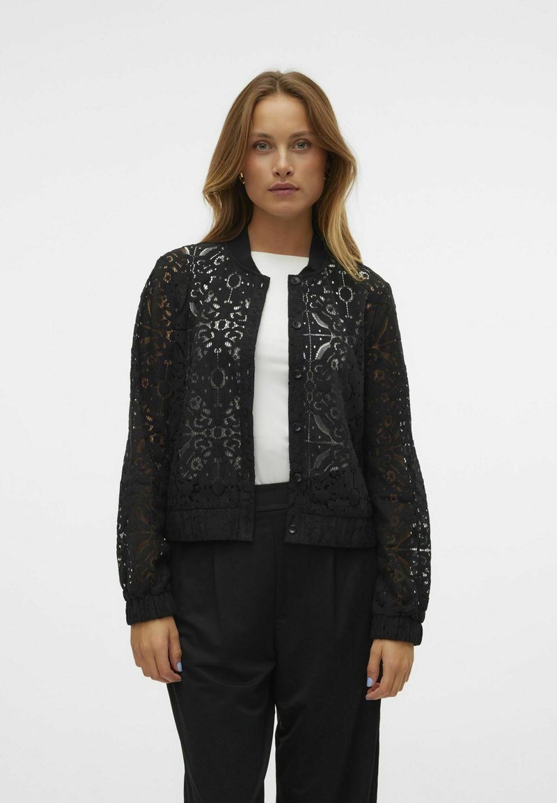 Veste en dentelle noire avec un col rond, fermeture à pression à l'avant et poignets côtelés. Présente des motifs floraux et géométriques sur l'ensemble. Portée sur un haut blanc.