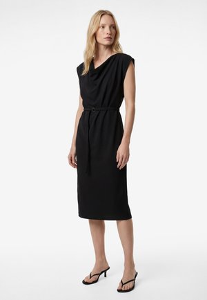 Vrouw draagt een mouwloze zwarte midi-jurk met een gedrapeerde halslijn en een dunne riem, gecombineerd met zwarte sandalen met hak, staand op een witte achtergrond.