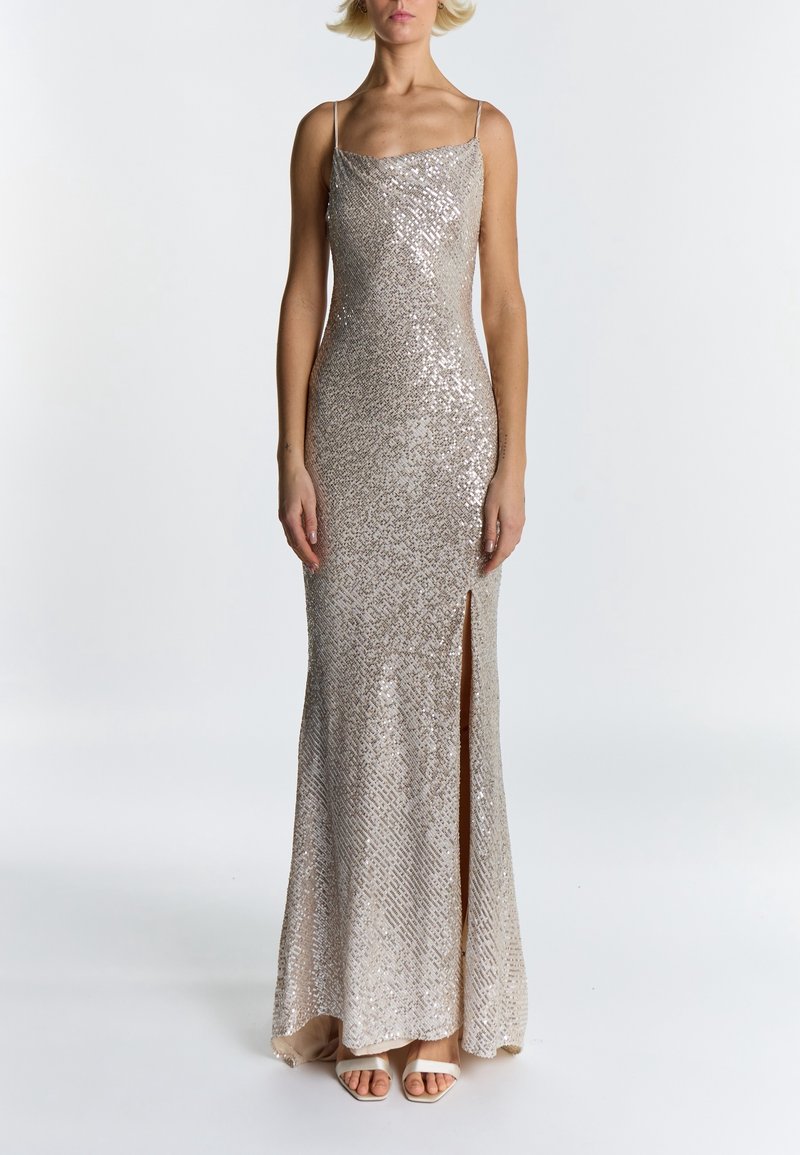 Femme portant une robe sleeveless argentée à sequins avec une fente devant et des sandales blanches à talons ouverts, debout sur un fond blanc uni.