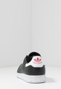 Svart läder sneaker med vit sula, med perforeringar och en rosa accent på hälfliken; "stan smith"-logotypen visas framträdande.