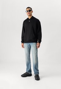 Moletom preto com bolso frontal, jeans claros desbotados e tênis pretos. Modelo usando óculos de sol, posando em frente a um fundo simples.